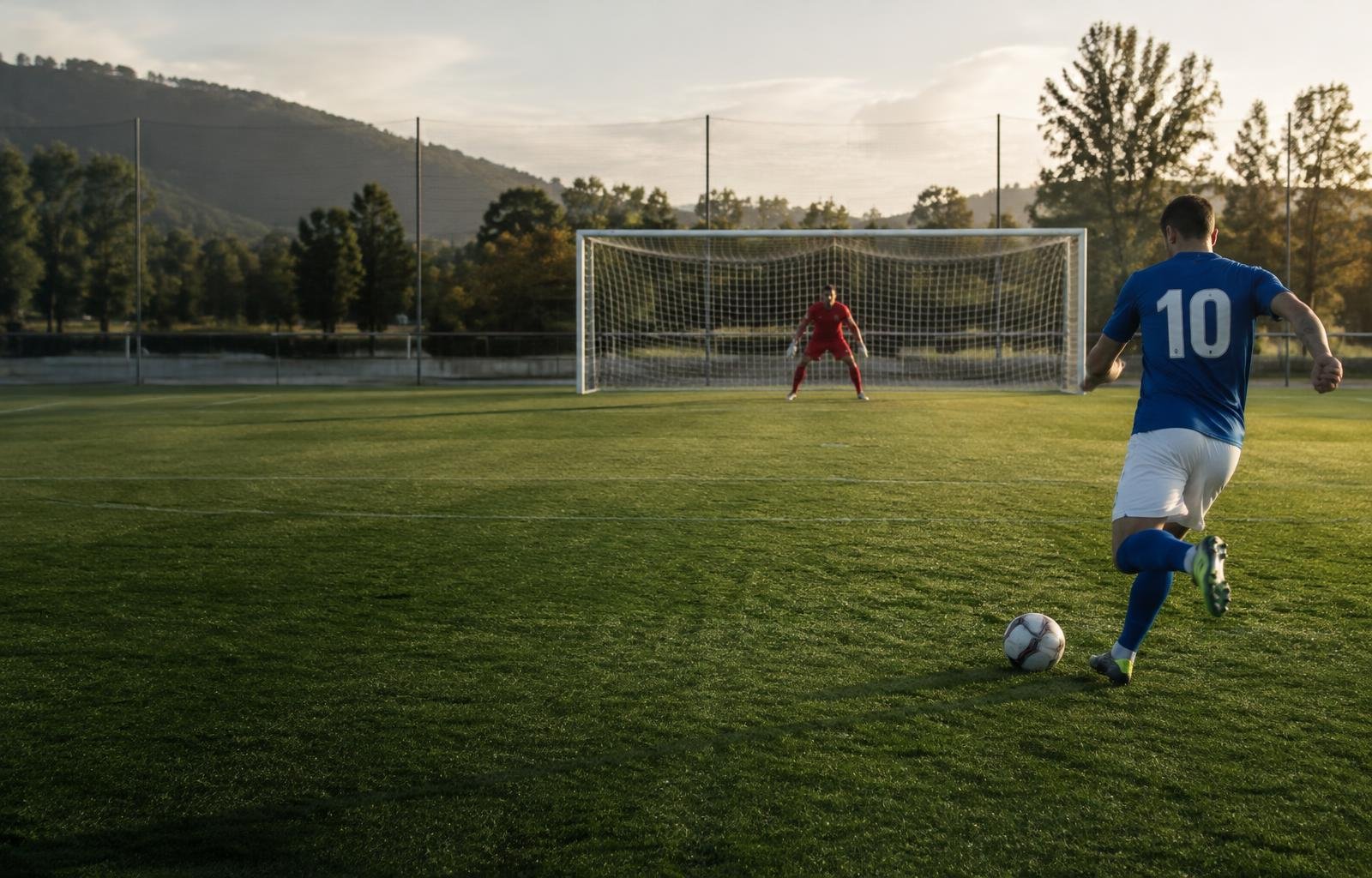 Batedor diante da bola e goleiro posicionado no gol em um campo de futebol